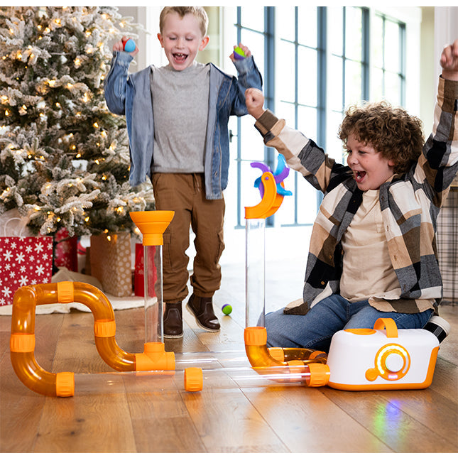 Two boys play with air-powered clear and orange tubes, a white base, and a spinning blue fan to launch balls.