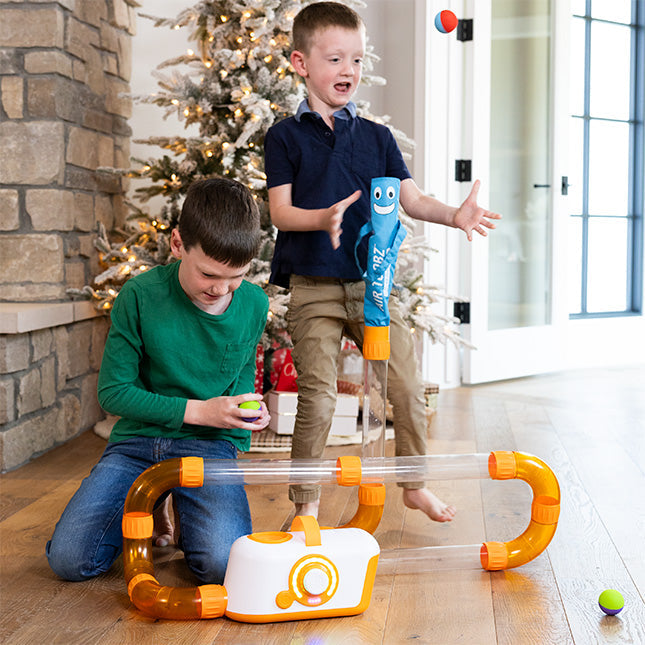 Two boys play with an air-powered toy featuring clear tubes, orange connectors, and a blue inflatable tube character.