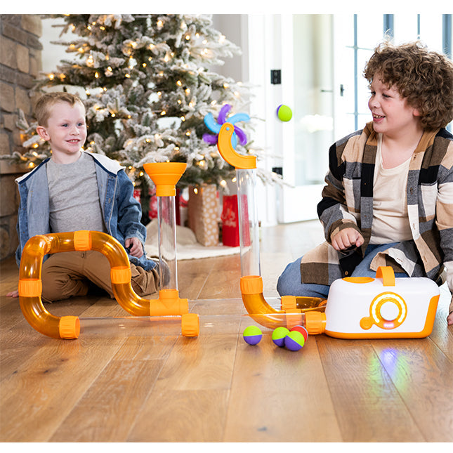 Two boys play with orange and clear air tubes, colorful balls, and a purple spinning fan accessory on a wooden floor.