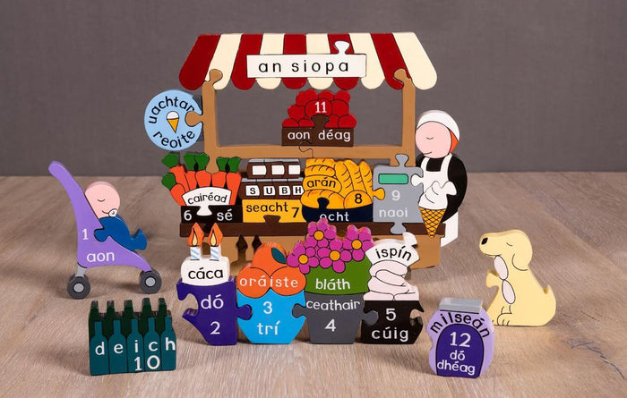 Colorful wooden puzzle of a shop with numbered pieces labeled in Irish, a shopkeeper figure, and a yellow dog.