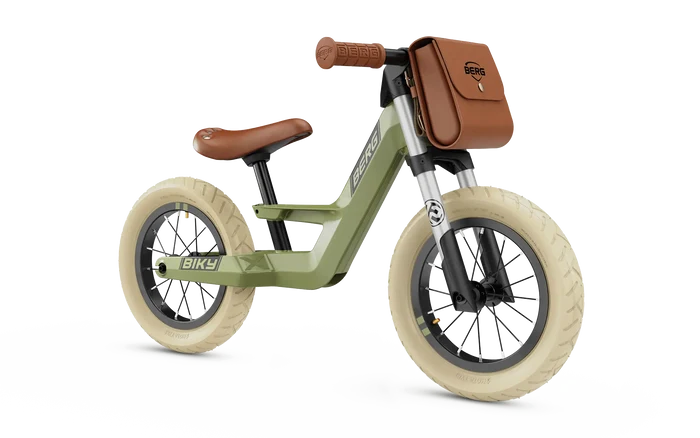 Sage green balance bike with cream tires, brown leather-look saddle, grips, and a matching front storage bag.