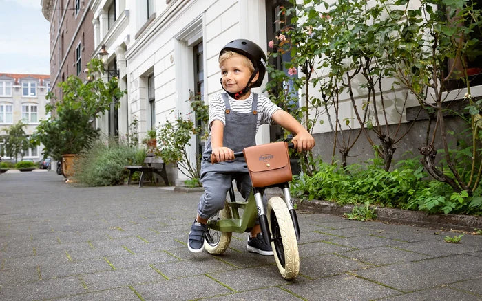 Boy on an olive green balance bike with cream tires and a brown handlebar bag on a paved sidewalk.