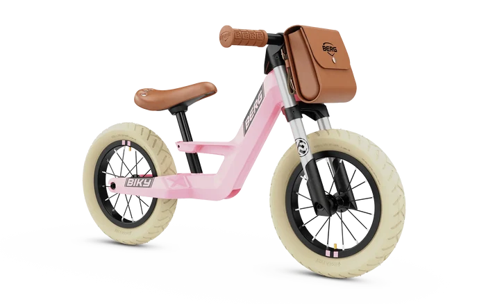 Pink retro balance bike with cream tires, brown leather seat, and a matching front-mounted handlebar bag.