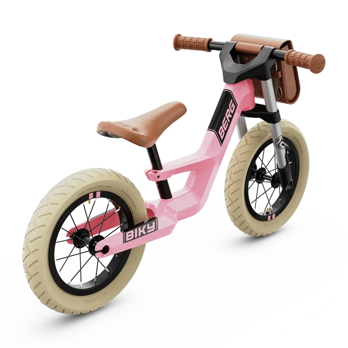Pink retro balance bike with cream tires, brown saddle and grips, and a small front handlebar bag.