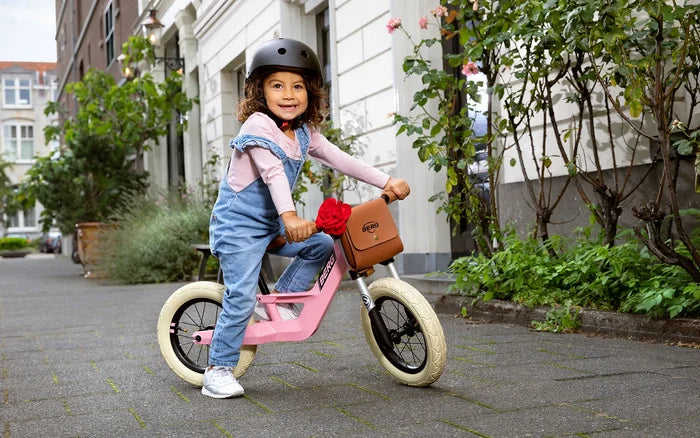 Child riding a pink retro-style balance bike with cream tires and a brown handlebar bag with a red flower.