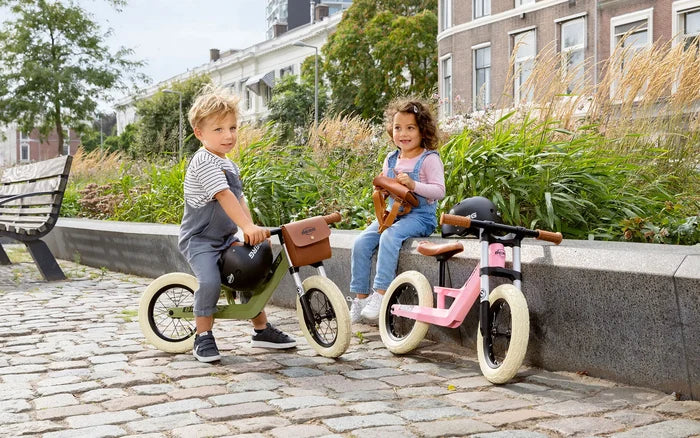 Pink retro balance bike with wide white tires and brown saddle, beside a girl sitting on a stone wall in a park.