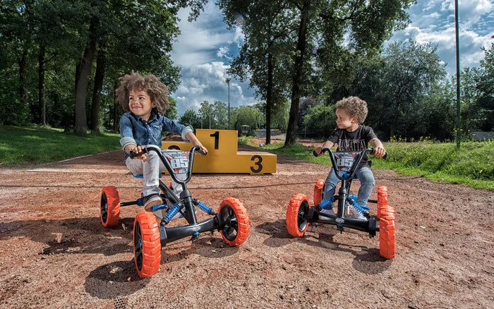 Two kids riding black off-road pedal go-karts with chunky orange tires and blue springs on a dirt track.