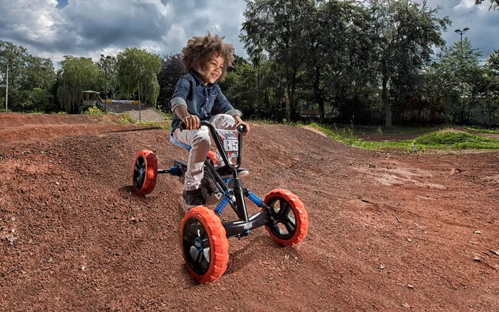 Child riding a black pedal go-kart with chunky orange tires and blue suspension springs on a dirt track.
