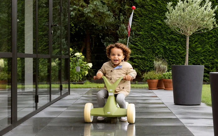 Child riding an olive green four-wheel pedal kart with cream wheels and a red flag on a stone patio.