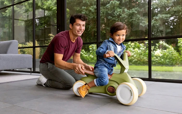 Child rides an olive green four-wheel toy with cream wheels and brown seat, assisted by an adult on a patio.