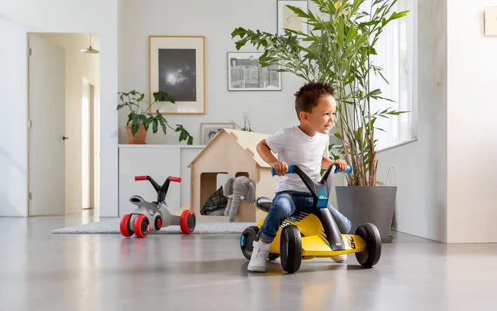 Child riding a yellow four-wheeled pedal kart with blue grips and black accents in a modern, sunlit indoor room.