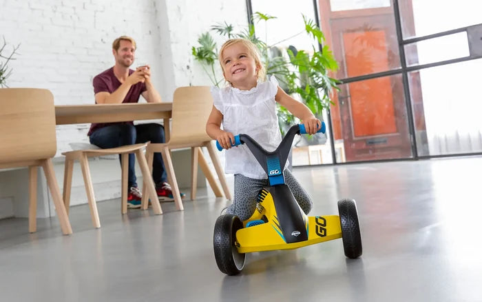 Toddler girl riding a yellow and black four-wheeled ride-on toy with blue handles in a modern home interior.
