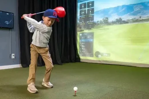 Child swinging an oversized red toy golf driver toward a white ball on a red tee in an indoor golf simulator.