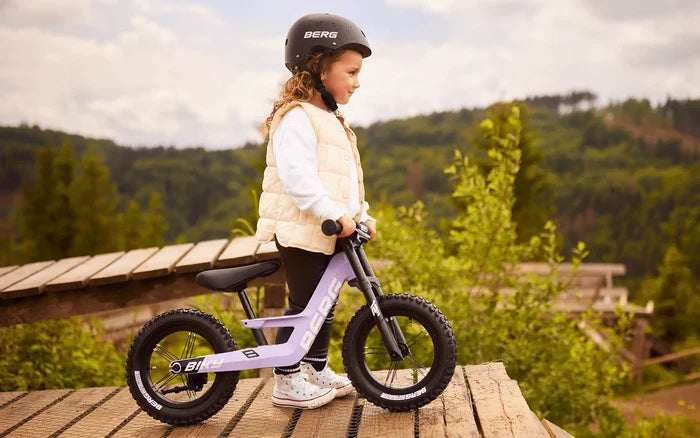 Child in a helmet with a purple balance bike featuring chunky off-road tires and a handbrake on a wooden platform.