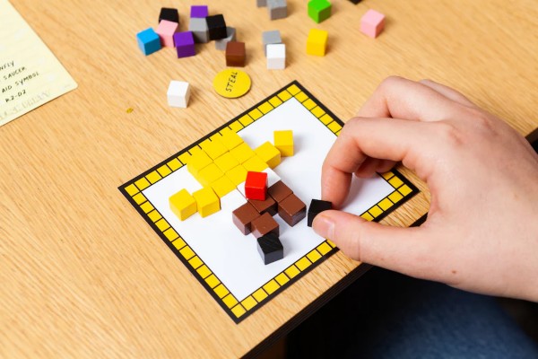 Hand building a pixel art character with tiny colored wooden blocks on a game board card.