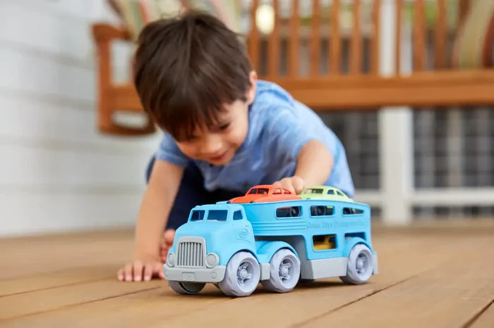 Boy playing with a blue toy car carrier truck carrying orange and green cars on a wooden deck.