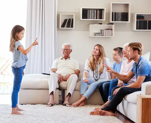 Multi-generational family laughing as a young girl acts out a clue during a game of charades in a bright living room.