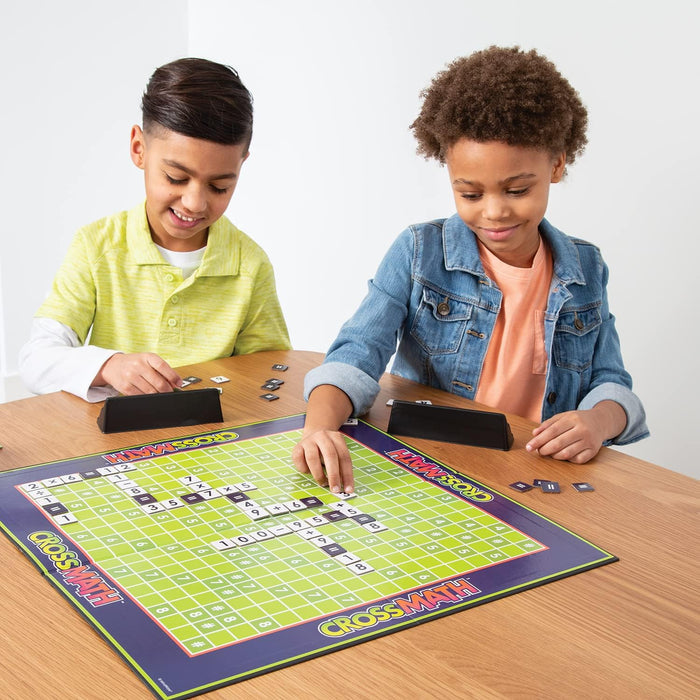 Two children playing a math crossword board game with number and symbol tiles on a green grid.