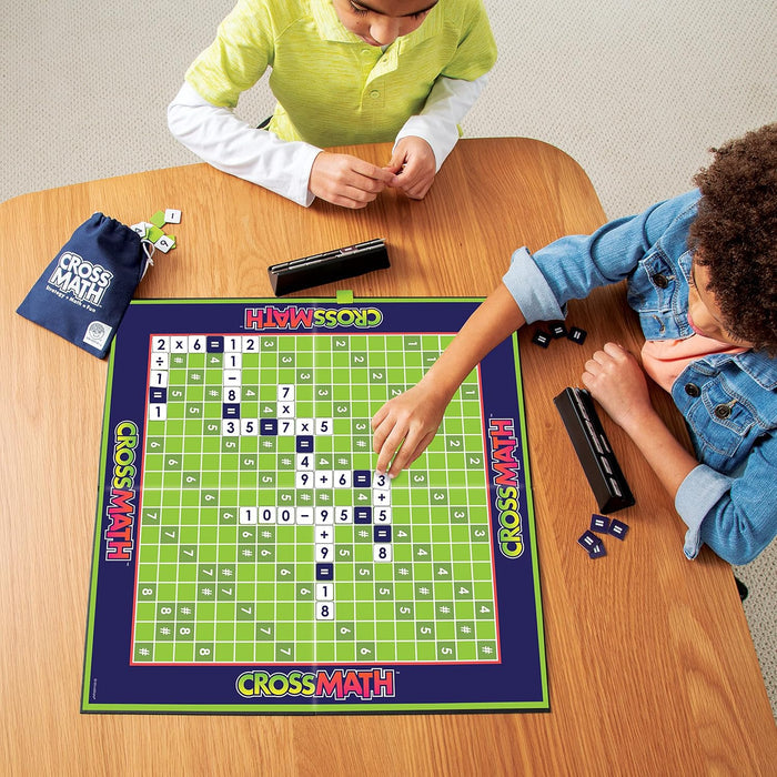 Two children play a math tile game on a green grid board with black racks and a blue drawstring bag.