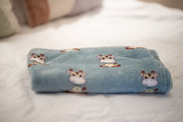 Folded soft blue plush blanket with a cartoon giraffe pattern on a white textured bedspread.