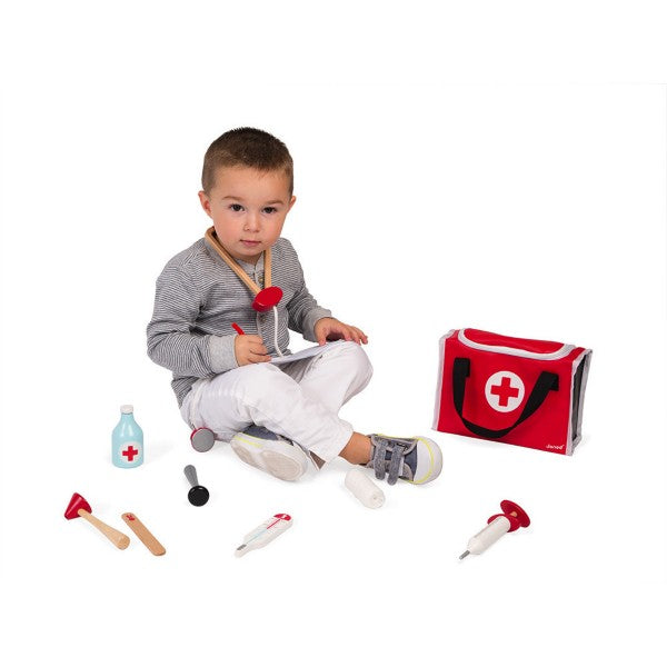 Toddler sitting with a red doctor's kit including a wooden stethoscope, syringe, and thermometer on a white background.
