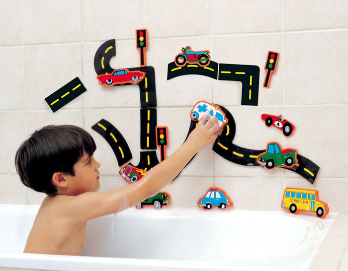 Child in a bathtub playing with colorful foam road tracks and vehicle bath toys stuck to the wall tiles.