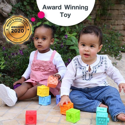 Two toddlers playing with colorful, textured square building blocks outdoors on a stone patio.