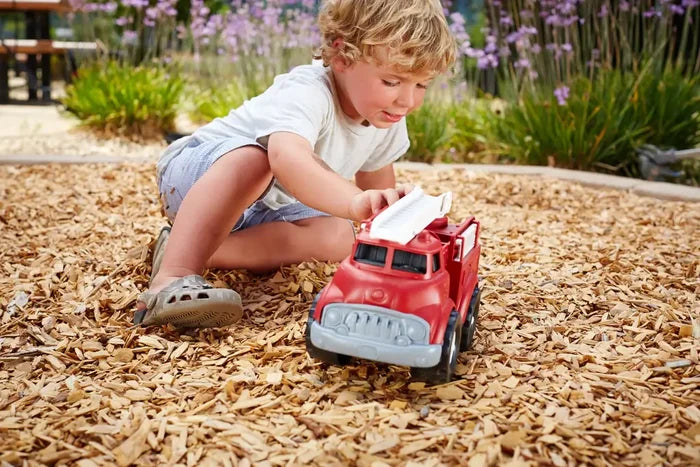 Toddler playing with a chunky red toy fire truck featuring a white ladder and grey grille on wood chips.