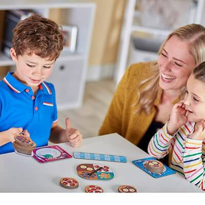 A smiling woman and two children playing a board game with pizza and pancake tokens and a spinner on a table.