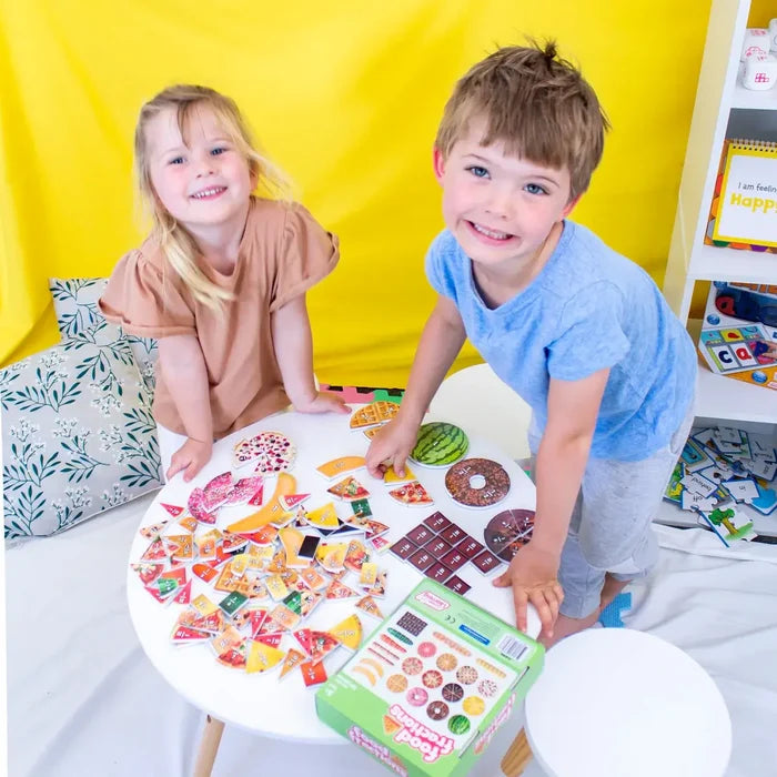 Two children playing with colorful food fraction puzzle pieces and a green box on a white table.