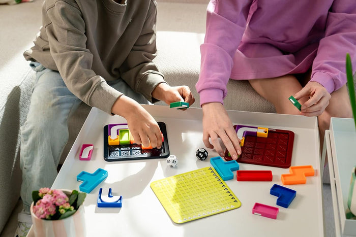 Hands placing colorful geometric blocks onto grid boards with dice and a yellow game card on a white table.