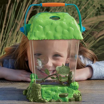 Clear plastic habitat with green textured lid and base, blue handle, and a girl smiling at a frog on a branch inside.