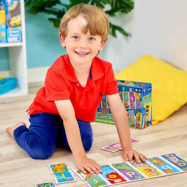 Boy in red shirt assembling long alphabet floor puzzle with colorful animals next to its box on a light wood floor.