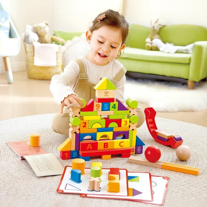 Young girl building a tower with colorful wooden blocks in various shapes, letters, and numbers on a soft rug.