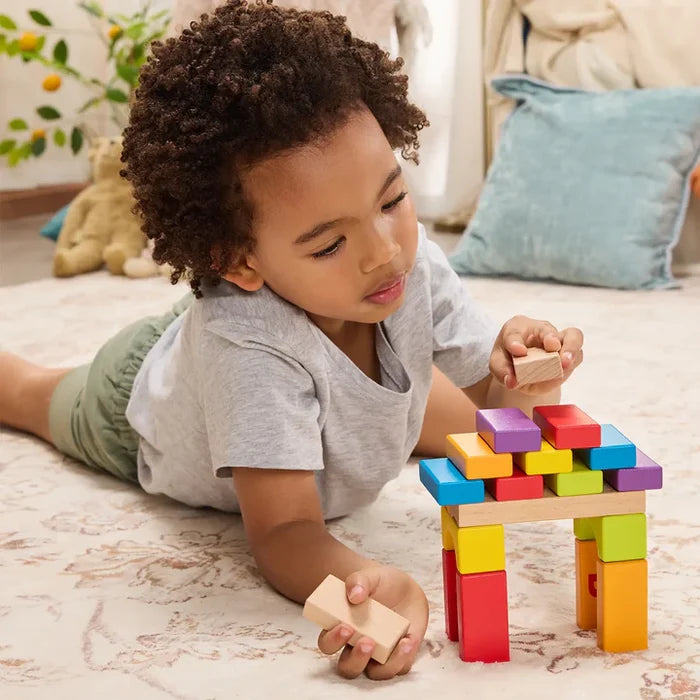Young child lying on a rug builds a colorful structure with rectangular and arched wooden blocks.