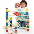 A child plays with a multi-level wooden marble run featuring colorful blocks, spiral funnels, and slanted tracks.