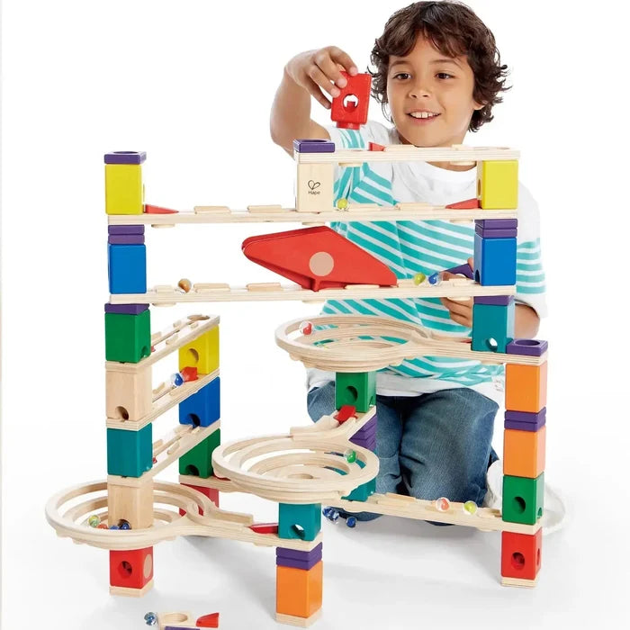 A child plays with a multi-level wooden marble run featuring colorful blocks, spiral funnels, and slanted tracks.