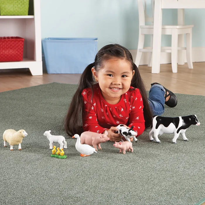 Smiling girl on a green rug plays with plastic toy cow, pig, sheep, and goose figurines with their babies.