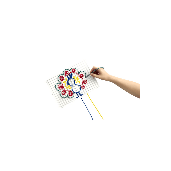 Hand threading green lace onto a white pegboard to create a colorful peacock with blue, red, and yellow strings.