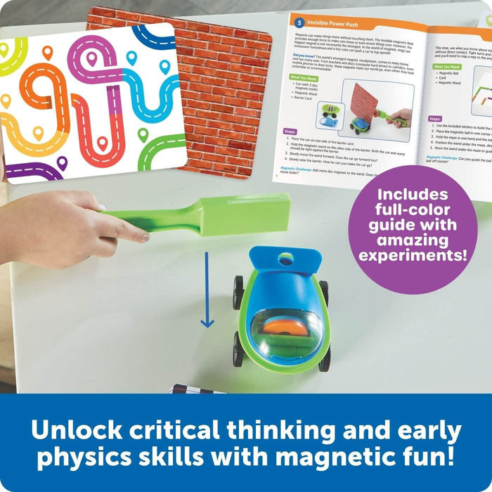 Hand holding a green magnetic wand near a blue toy car, with an open experiment guide and colorful activity cards.