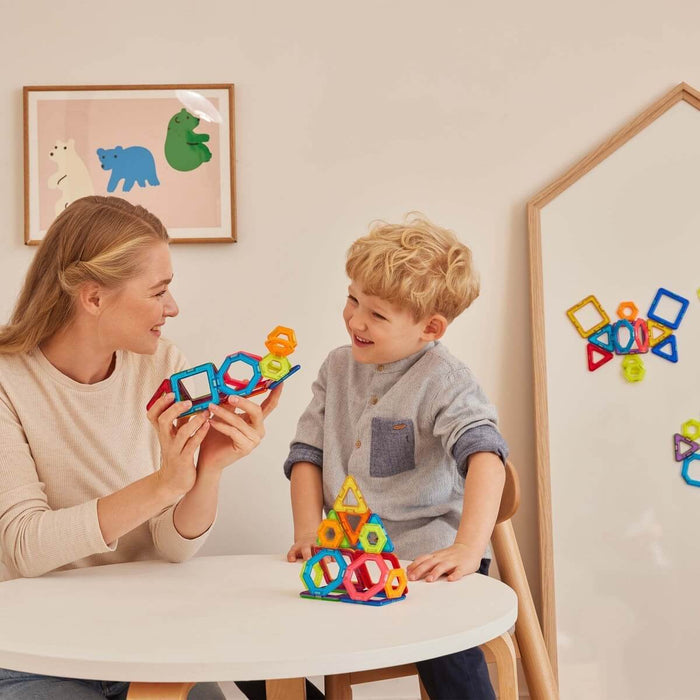 Woman and child building with colorful magnetic squares, triangles, and hexagons on a white table.