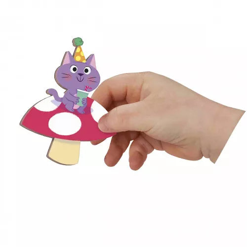 Hand holding a magnetic piece of a purple cat in a party hat sitting on a red and white spotted mushroom.
