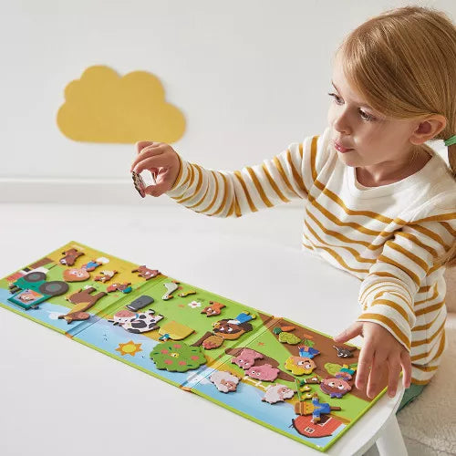 A young girl playing with a magnetic farm-themed fold-out scene featuring animals and characters on a white table.
