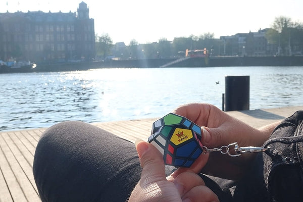 Mini black dodecahedron puzzle keychain with colorful stickers held in hands outdoors by a sunny waterfront.
