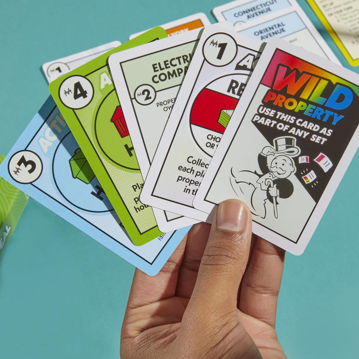 A hand holds a fan of Monopoly Deal cards, including property and wild cards, against a teal background.