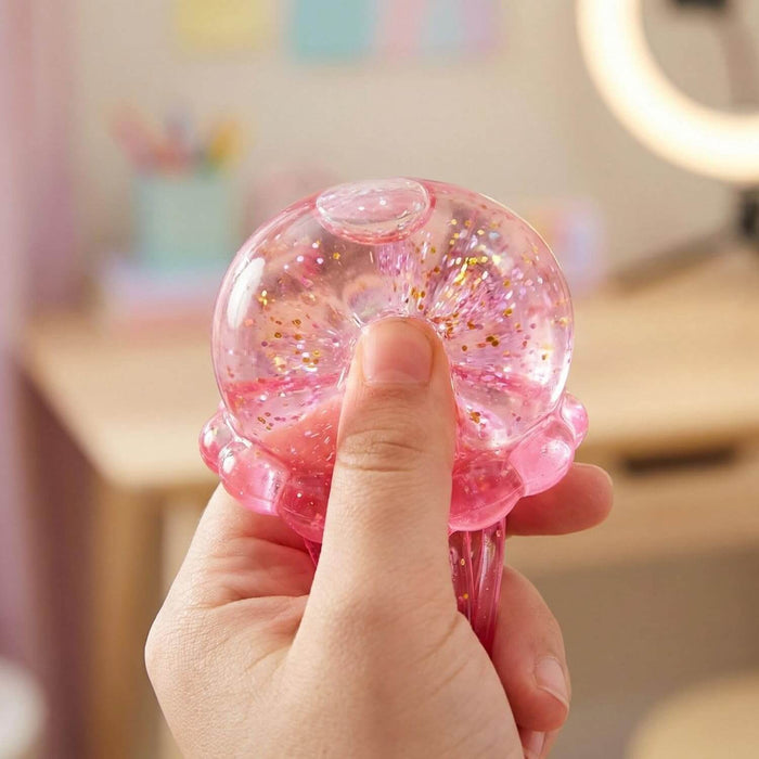 Hand squeezing a translucent pink jelly dome filled with gold glitter, showing its soft, squishy texture.