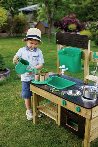 Wooden mud kitchen with green sink, water dispenser, and stove. A child waters small potted plants on the dark counter.