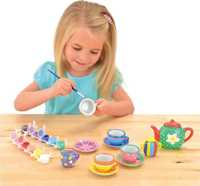 Girl painting a white ceramic cup next to a colorful floral teapot, cups, and paint pots on a wooden table.