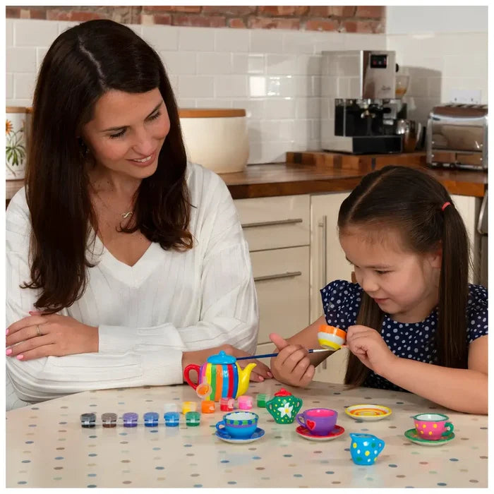 A child paints a miniature ceramic tea set with colorful stripes and patterns as a woman watches at a table.