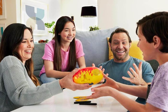 Family plays a game as a woman passes a yellow and red flame-patterned potato-shaped electronic toy to a boy.